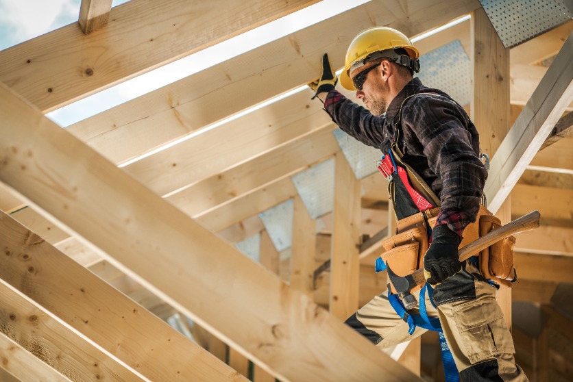 The image shows a construction worker wearing a hard hat and tool belt, working on a building with wooden beams. This image pairs with the article on reasons why workers' compensation is denied.