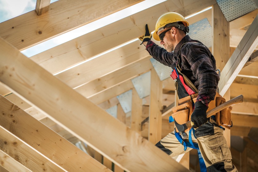 The image shows a construction worker with a hard hat wearing a tool belt, while working on a building with wooden beams.